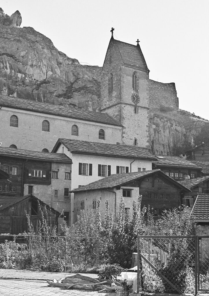 pexels-photo-29229353-29229353 Black and white photograph of a historic stone church surrounded by village houses against a rocky backdrop.